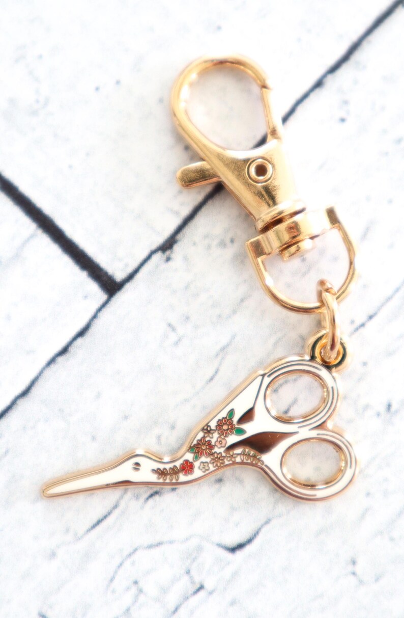 May include: Gold-tone metal key chain with a floral patterned scissor charm. The scissor charm has a white background with gold-tone metal accents and a floral pattern in shades of red, green, and brown.