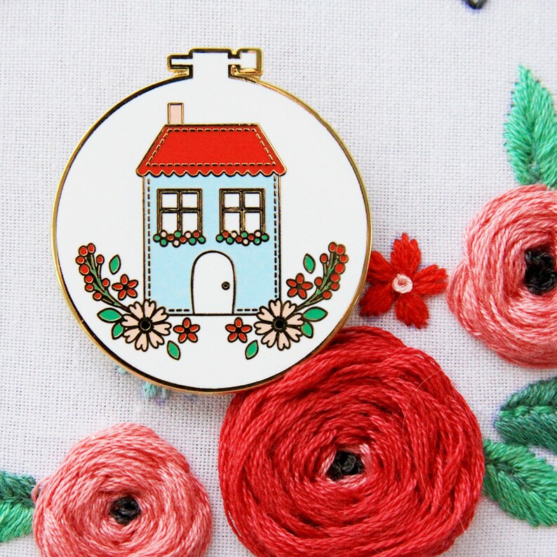 K&ouml;nnte beinhalten: Ein blaues Haus mit rotem Dach und wei&szlig;en Verzierungen ist von Blumen in einem wei&szlig;en Stickrahmen umgeben. Der Rahmen ist goldfarben und hat oben eine kleine Schlaufe.