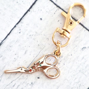 May include: Gold-tone key chain with a floral patterned scissor charm. The scissor charm has a long, thin blade and a rounded handle.