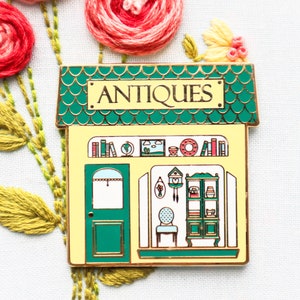 May include: A gold-trimmed enamel pin in the shape of an antique shop. The shop has a green roof and a yellow facade with the word "ANTIQUES" above the door. The shop's interior is visible, with shelves, a chair, and various decorative items.