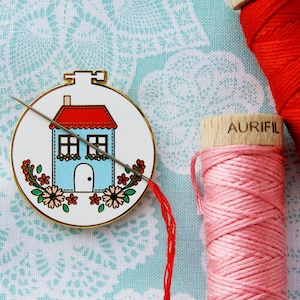 K&ouml;nnte beinhalten: Ein blaues und rotes Haus mit einer wei&szlig;en T&uuml;r und Fenstern ist auf einem wei&szlig;en runden Reifen mit goldener Einfassung gestickt. Eine Nadel und roter Faden sind durch den Reifen gef&auml;delt. Zwei Garnrollen, eine rote und eine rosafarbene, befinden sich im Hintergrund.