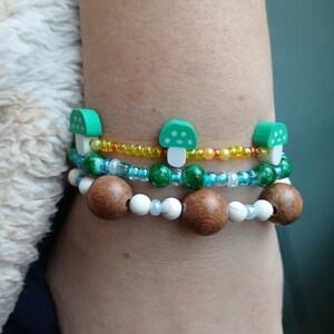 May include: A stack of three bracelets with green mushroom charms, white beads, and brown wooden beads.