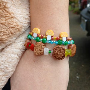 May include: A colorful beaded bracelet with wooden mushroom charms and yellow mushroom beads. The bracelet is on a person's wrist.