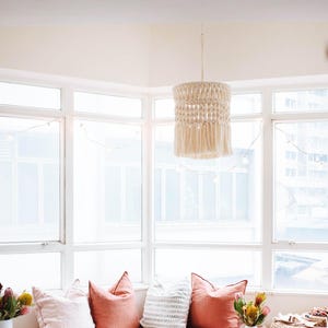 Op de afbeelding: Een gezellige vensterbank met koraal- en blushkleurige kussens, een patroonkleed en een roze deken. Boven hangt een macramé hanglamp. Decoratieve vazen en kleine houten tafels met drankjes en bloemen maken de scène compleet.