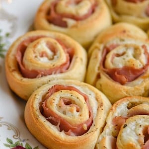 May include: Close-up of several baked ham and cheese rolls. The rolls have a golden-brown exterior and a spiral of ham and cheese filling. The rolls are arranged on a white plate.
