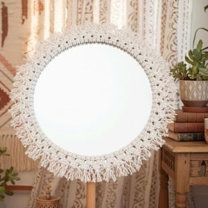 May include: A circular mirror with a white macrame fringe. The mirror is supported by a light-coloured wooden stand. The decorative mirror is a bohemian style home decor item.