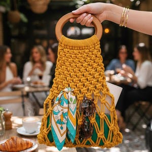 Peut inclure: Sac &agrave; main en macram&eacute; jaune moutarde avec une poign&eacute;e en bois. Le sac contient une &eacute;charpe &agrave; motifs, des lunettes de soleil et une pochette verte. Le sac est tenu par une personne avec des bracelets dor&eacute;s. Sc&egrave;ne de caf&eacute; en arri&egrave;re-plan.