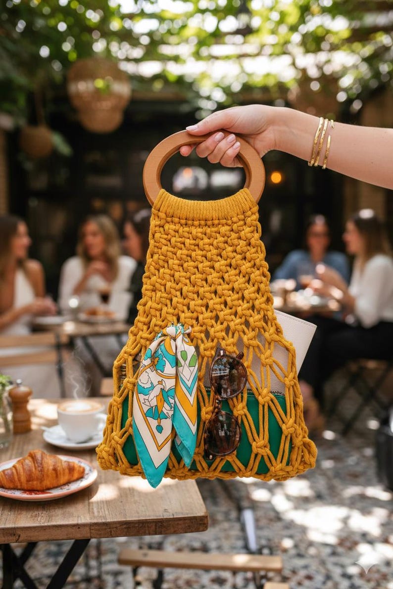 Peut inclure: Sac &agrave; main en macram&eacute; jaune moutarde avec une poign&eacute;e en bois. Le sac contient une &eacute;charpe &agrave; motifs, des lunettes de soleil et une pochette verte. Le sac est tenu par une personne avec des bracelets dor&eacute;s. Sc&egrave;ne de caf&eacute; en arri&egrave;re-plan.