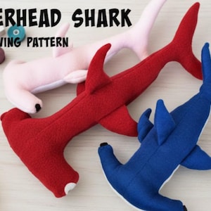 Puede incluir: Tres tiburones martillo de peluche en rojo, rosa y azul. La imagen incluye suministros de costura y el texto "HAMMERHEAD SHARK PLUSHIE SEWING PATTERN". También se ve una cinta métrica amarilla y una cinta métrica blanca.