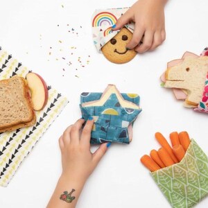 Op de afbeelding: Een overzichtsfoto toont diverse voedingsmiddelen verpakt in kleurrijke, herbruikbare bijenwasverpakkingen. Een sandwich, koekje en wortels zijn zichtbaar. De verpakkingen hebben patronen zoals regenbogen, bloemen en geometrische vormen.