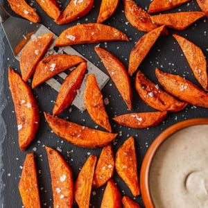 May include: A top-down view of baked sweet potato wedges, seasoned and salted, on a dark surface. A metal spatula is among the wedges. A small bowl of dipping sauce is in the bottom right corner. The wedges are a bright orange colour.