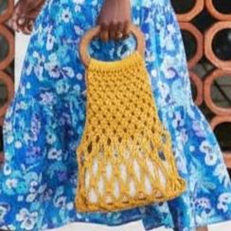 Peut inclure: Un sac en crochet jaune moutarde avec une poign&eacute;e en bois. Le sac pr&eacute;sente un motif ajour&eacute; et une doublure blanche. Le sac est tenu par une personne portant une robe bleue &agrave; fleurs.
