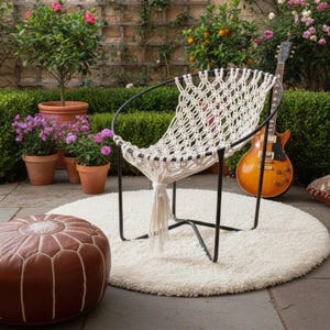 May include: A white macrame chair with a black metal frame sits on a white round rug. A brown leather ottoman and a guitar are nearby. The chair is in a garden setting with potted plants and flowers.