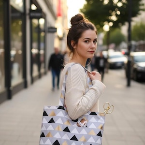 Puede incluir: Un bolso tote con un patrón geométrico de triángulos negros, dorados y grises. El bolso tiene un fondo blanco y asas negras. Se ven unas tijeras doradas y una cinta métrica. La persona lleva un suéter color crema.