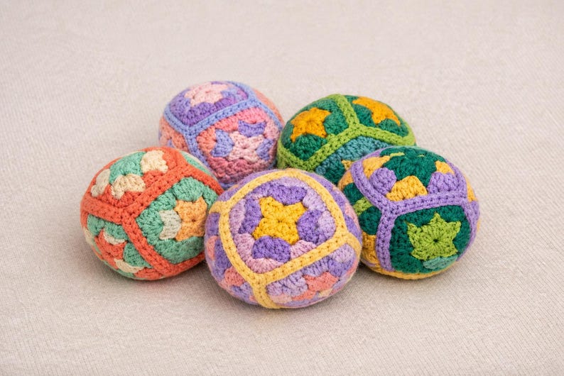 Puede incluir: Cinco bolas de ganchillo coloridas en tonos naranja, verde, morado y amarillo. Cada bola presenta un patr&oacute;n geom&eacute;trico &uacute;nico. Las bolas est&aacute;n dispuestas sobre una superficie neutra y texturizada.