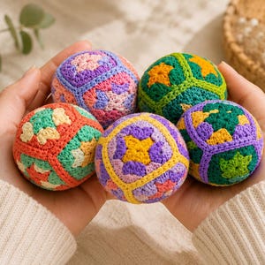 Puede incluir: Primer plano de cinco bolas de ganchillo coloridas con patrones geom&eacute;tricos, sostenidas en manos abiertas. Las bolas presentan tonos de rosa, morado, verde, naranja y amarillo. El fondo es de un color suave y neutro, resaltando los art&iacute;culos hechos a mano.