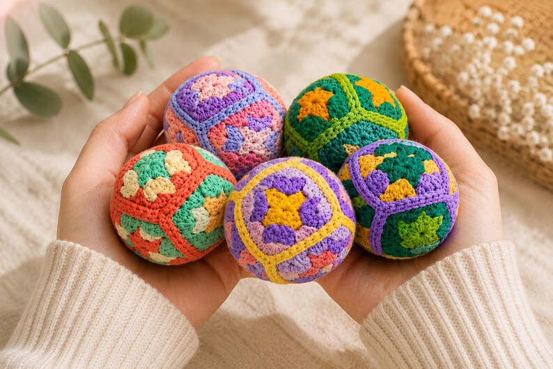 Puede incluir: Primer plano de cinco bolas de ganchillo coloridas con patrones geom&eacute;tricos, sostenidas en manos abiertas. Las bolas presentan tonos de rosa, morado, verde, naranja y amarillo. El fondo es de un color suave y neutro, resaltando los art&iacute;culos hechos a mano.