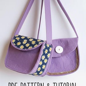 May include: Two small, lavender felt purses with long straps hang against a white background. One purse has a flap with a navy blue and white daisy pattern. The other has a white button closure. Text at the bottom reads "PDF PATTERN & TUTORIAL".