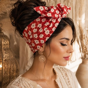 May include: A red headband with a white floral pattern is tied in a bow. The headband is worn on the head. The person is wearing gold earrings and a white lace top. The background is blurred.