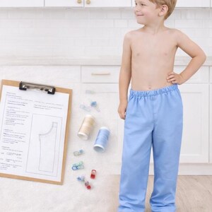 May include: A young child wearing light blue pajama pants. Sewing supplies, including thread spools and clips, are next to a clipboard with a pattern. The child is standing in front of a white kitchen.