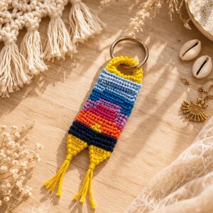 Op de afbeelding: Een handgemaakte macram&eacute; sleutelhanger met een kleurrijk zonsondergangontwerp. De sleutelhanger heeft een gele bovenkant en kwastjes, die overgaan in tinten blauw, roze en oranje. Een goudkleurige ring is bevestigd voor sleutels.