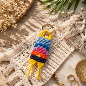 Op de afbeelding: Een handgemaakte, kleurrijke sleutelhanger met een gouden ring. De sleutelhanger heeft een geweven ontwerp met blauwe, roze, oranje en gele tinten. De sleutelhanger wordt gepresenteerd op een macrame placemat.