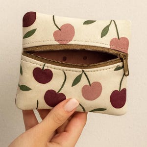 May include: A small, rectangular pouch with a cream-colored background and a repeating pattern of cherries in shades of pink and burgundy, with green leaves. The pouch has a brown zipper and is partially open, revealing a brown interior.