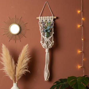 Op de afbeelding: Een crèmekleurige macramé plantenhouder met een glazen pot met groen. De houder is opgehangen aan een houten deuvel en heeft een geknoopt ontwerp met een lange kwast. De achtergrond is een warme bruine muur.