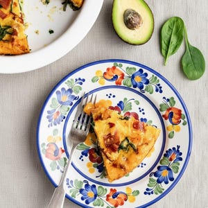 Könnte beinhalten: Ein Stück Quiche mit Speck und Spinat auf einem dekorativen Teller mit blauen, orangefarbenen und gelben Blumenmustern. Eine Gabel liegt neben der Quiche. Eine halbe Avocado und Spinatblätter sind ebenfalls zu sehen.