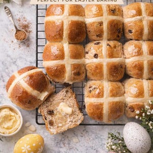 May include: Overhead shot of freshly baked hot cross buns arranged on a wire rack. The buns are golden brown with a white cross on top. A slice of bread with butter, a bowl of butter, and decorated Easter eggs are also visible.