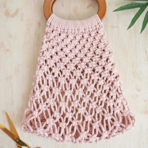 Peut inclure: Un sac fourre-tout en macram&eacute; rose clair avec une poign&eacute;e en bois. Le sac pr&eacute;sente un motif tiss&eacute; et une poign&eacute;e arrondie. Des ciseaux dor&eacute;s sont visibles dans le coin inf&eacute;rieur gauche. Le sac est sur une surface en bois clair.