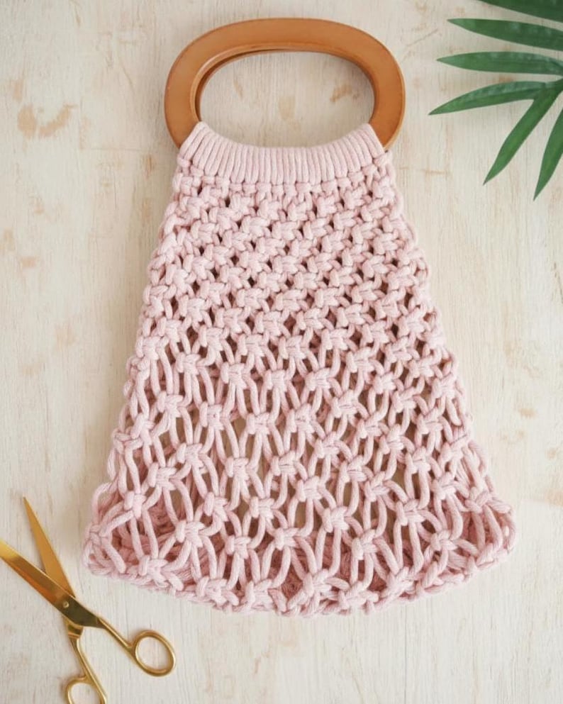 Peut inclure: Un sac fourre-tout en macram&eacute; rose clair avec une poign&eacute;e en bois. Le sac pr&eacute;sente un motif tiss&eacute; et une poign&eacute;e arrondie. Des ciseaux dor&eacute;s sont visibles dans le coin inf&eacute;rieur gauche. Le sac est sur une surface en bois clair.