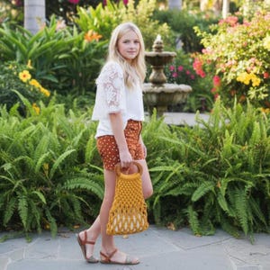 Peut inclure: Une jeune personne porte un haut en dentelle blanche et un short &agrave; pois marron, tenant un sac en macram&eacute; jaune moutarde avec des poign&eacute;es en bois. Le sac mesure environ 30 cm de haut. Le fond pr&eacute;sente une v&eacute;g&eacute;tation luxuriante et une fontaine.