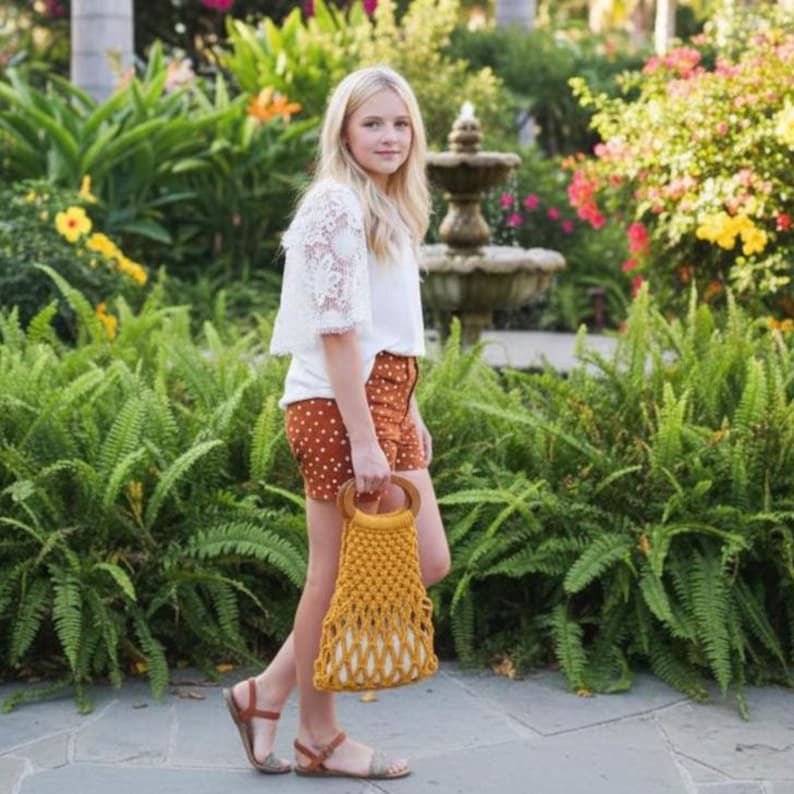 Peut inclure: Une jeune personne porte un haut en dentelle blanche et un short &agrave; pois marron, tenant un sac en macram&eacute; jaune moutarde avec des poign&eacute;es en bois. Le sac mesure environ 30 cm de haut. Le fond pr&eacute;sente une v&eacute;g&eacute;tation luxuriante et une fontaine.