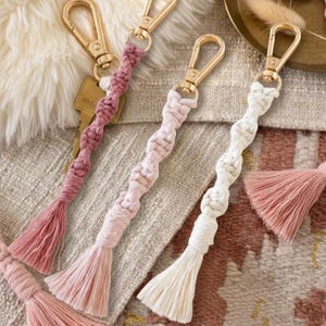Könnte beinhalten: Nahaufnahme mehrerer Makramee-Schlüsselanhänger mit goldfarbenen Verschlüssen und Quasten. Die Schlüsselanhänger sind in Rosa- und Weißtönen gehalten und haben ein gedrehtes Seil-Design. Die Quasten bestehen aus weichem, gefranstem Material. An einem Schlüsselanhänger ist ein Schlüssel befestigt.