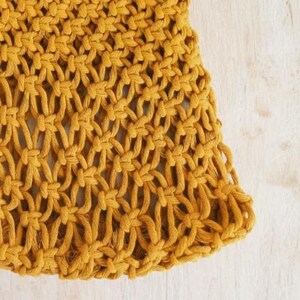 Peut inclure: Gros plan sur une section d'un textile macram&eacute; jaune moutarde. Le motif nou&eacute; cr&eacute;e un design textur&eacute; et ajour&eacute;. Le textile est pos&eacute; sur un fond clair en bois, mettant en valeur la couleur et la texture du macram&eacute;.