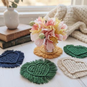 May include: A collection of heart-shaped macrame coasters in various colors, including navy blue, green, and cream. A bouquet of pink, yellow, and white paper flowers sits in a small glass jar. A yellow coaster is under the jar.