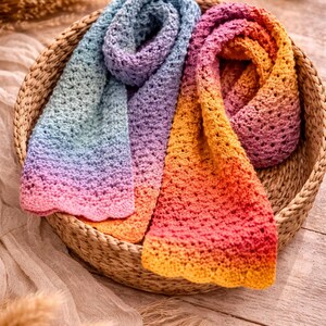 May include: Two crocheted scarves in a woven basket. One scarf fades from blue to pink, the other from orange to pink. The scarves are made of soft yarn and have a textured appearance. The basket sits on a wooden surface.