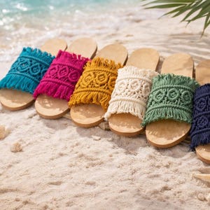 Op de afbeelding: Een rij kleurrijke macramé sandalen in tinten turquoise, fuchsia, mosterdgeel, crème, groen en marineblauw. De sandalen zijn geplaatst op een zandstrand met schelpen en een zeester.