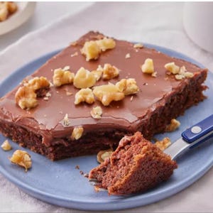 以下が含まれることがあります： 光沢のあるチョコレートフロスティングと刻んだクルミをのせたチョコレートケーキのスライスが、水色のプレートにのっています。ケーキの一部がフォークにのっています。クルミの小さなボウルが背景にあります。
