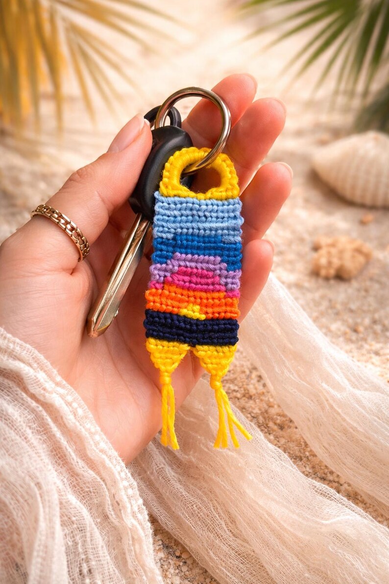 Op de afbeelding: Een kleurrijke, handgemaakte sleutelhanger met een zonsondergangontwerp. De sleutelhanger heeft een geweven patroon in tinten geel, blauw, paars en oranje. Het is bevestigd aan een zilveren sleutelring met autosleutels.