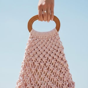 May include: A light pink macrame handbag with a wooden handle. The bag is held up against a clear blue sky. The bag's woven design is visible, and the wooden handle is a light brown color.