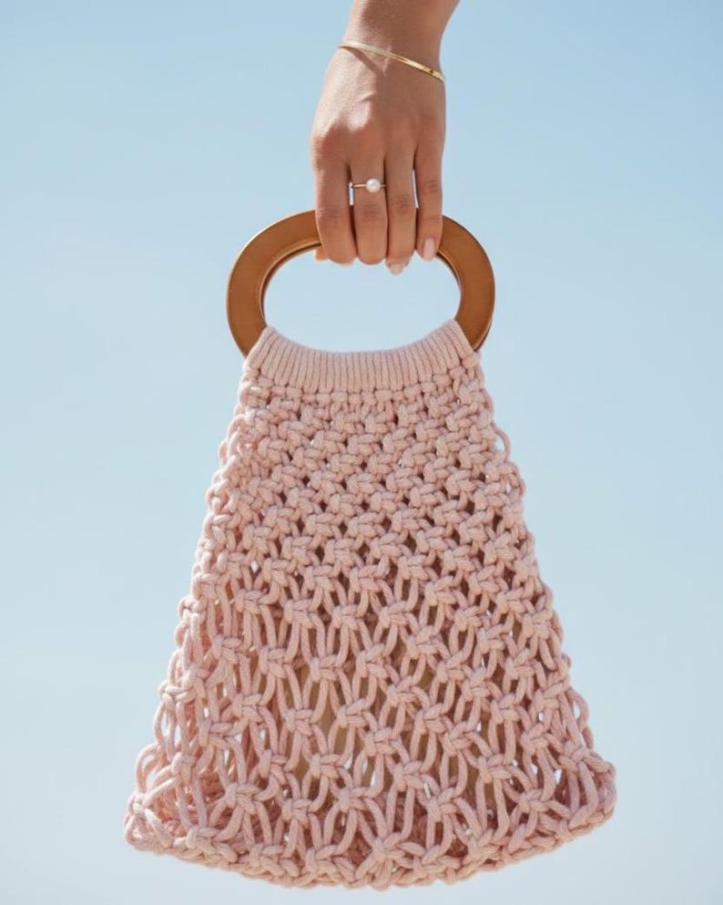 Peut inclure: Un sac &agrave; main en macram&eacute; rose clair avec une poign&eacute;e en bois. Le sac est tenu contre un ciel bleu clair. Le design tiss&eacute; du sac est visible, et la poign&eacute;e en bois est de couleur marron clair.