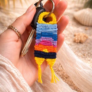 Op de afbeelding: Een kleurrijke, handgemaakte sleutelhanger met een zonsondergangontwerp. De sleutelhanger heeft een geweven patroon in tinten geel, blauw, paars en oranje. Het is bevestigd aan een zilveren sleutelring met autosleutels.