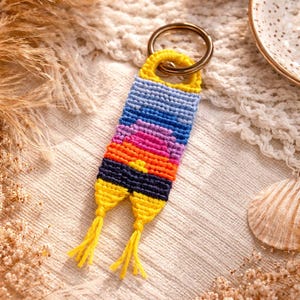 Op de afbeelding: Een handgemaakte sleutelhanger met een kleurrijk geweven ontwerp. De sleutelhanger heeft een kleurovergang van blauw, roze, oranje en geel, met een goudkleurige ring. De kwastjes zijn geel. De achtergrond is een neutraal gekleurde stof.