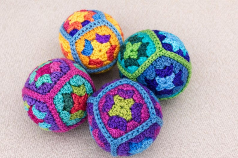 Puede incluir: Cuatro bolas de ganchillo coloridas en tonos morado, rosa, verde, azul, amarillo y naranja. Cada bola tiene un patr&oacute;n geom&eacute;trico y est&aacute; hecha con hilo. Las bolas est&aacute;n dispuestas sobre una superficie neutra y texturizada.