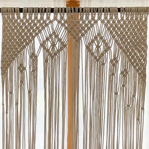 May include: A tan and beige macrame wall hanging with a detailed geometric design. The piece has long, vertical fringe and is suspended from a dark rod. This handcrafted item offers a bohemian style.