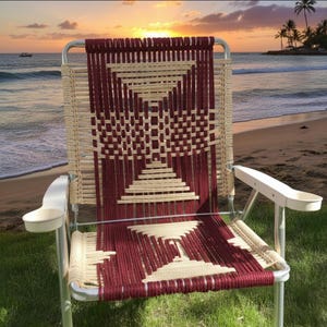 以下が含まれることがあります： シルバーフレームの、バーガンディとベージュの織りビーチチェア。カップホルダー付きの白いアームレストが付いています。背景には、海とヤシの木に沈む夕日のビーチシーンが写っています。