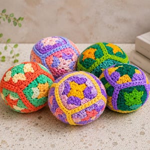 Puede incluir: Cinco bolas de ganchillo coloridas, en tonos naranja, verde, morado y amarillo, dispuestas sobre una superficie clara. Las bolas tienen patrones geom&eacute;tricos y est&aacute;n hechas a mano. Una planta en una maceta blanca est&aacute; al fondo.