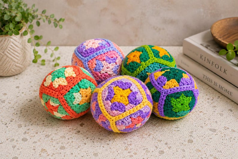 Puede incluir: Cinco bolas de ganchillo coloridas, en tonos naranja, verde, morado y amarillo, dispuestas sobre una superficie clara. Las bolas tienen patrones geom&eacute;tricos y est&aacute;n hechas a mano. Una planta en una maceta blanca est&aacute; al fondo.